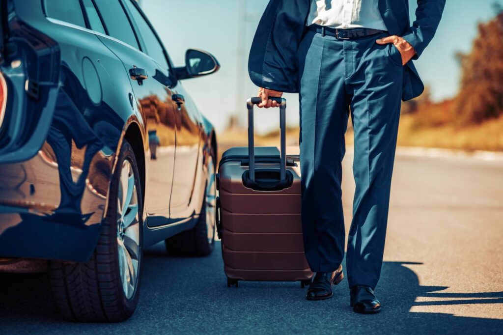 Como economizar combustível em viagens longas 5 Vendeu o carro e vai viajar - bagageiro de teto carro homem carregando mala de rodinhas perto de um carro